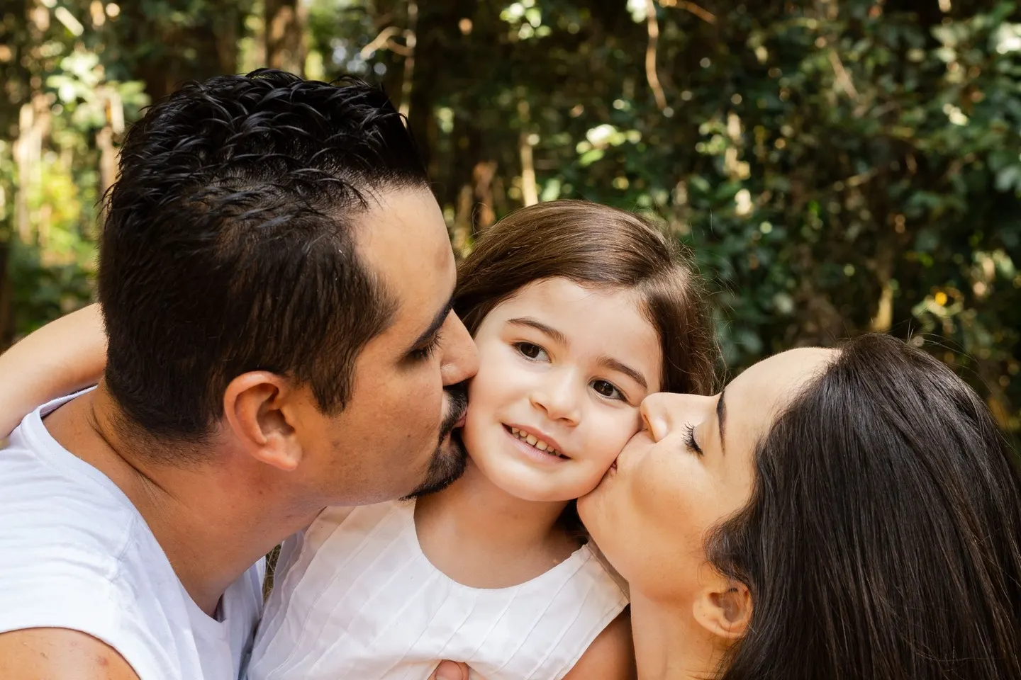 Pai e mãe beijando o rosto da filha pequena, que sorri