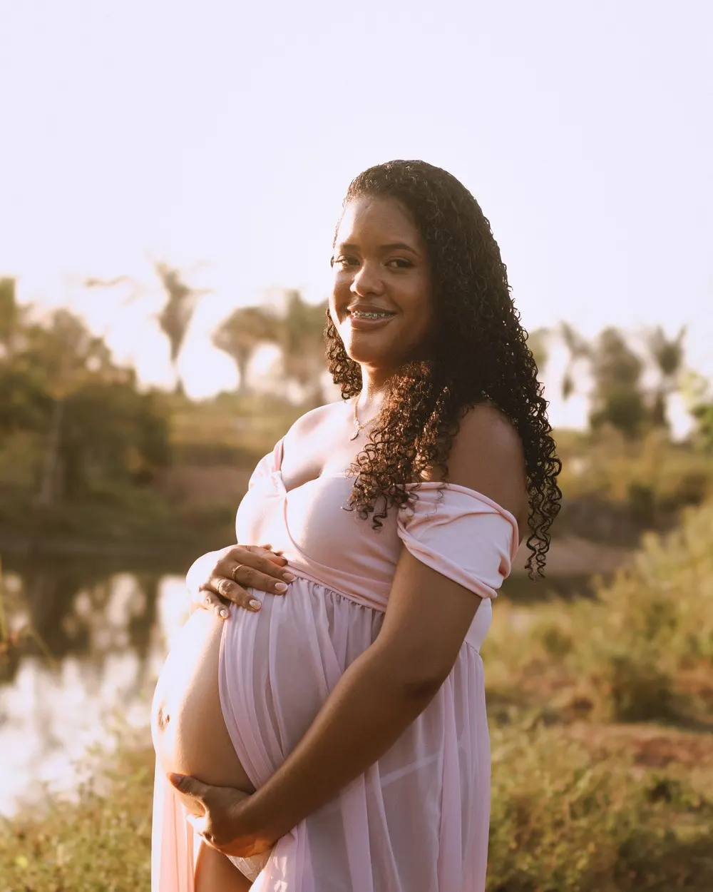 Gestante sorridente de vestido rosa ao ar livre, com as mãos na barriga