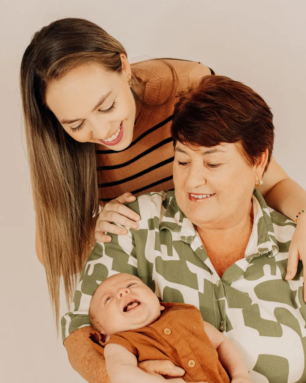 Três gerações: mulher jovem abraçando a mãe, que segura um bebê no colo, todos sorrindo