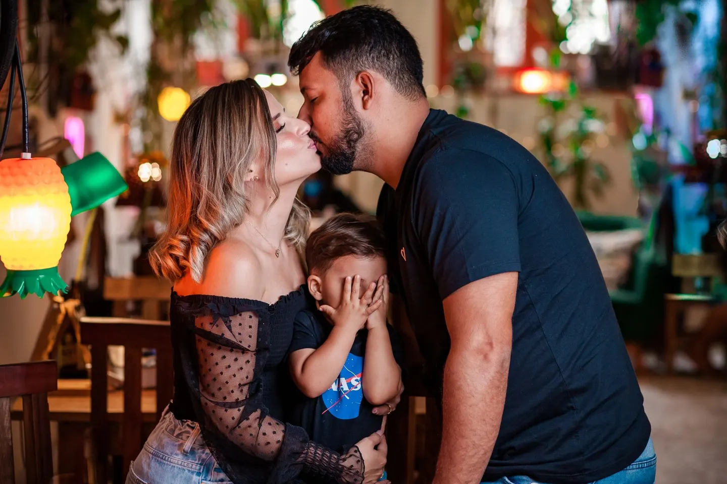 Casal se beijando enquanto o filho pequeno tapa os olhos, em casa